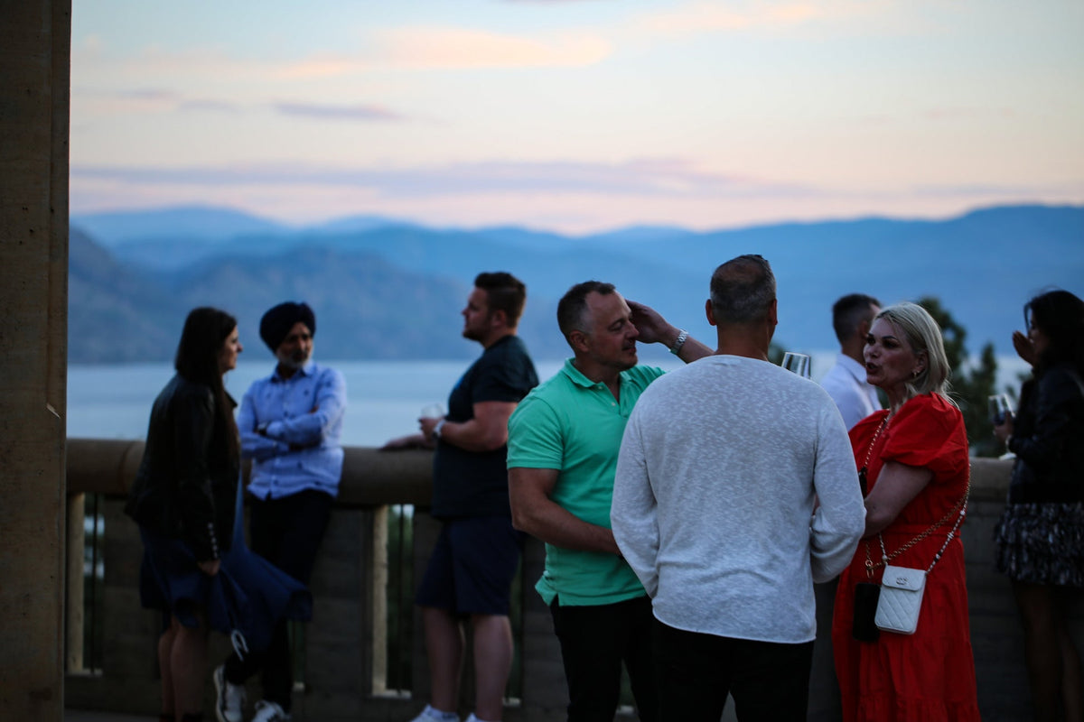 Past attendees talking over glasses of wine on a lakeside terrace at dusk